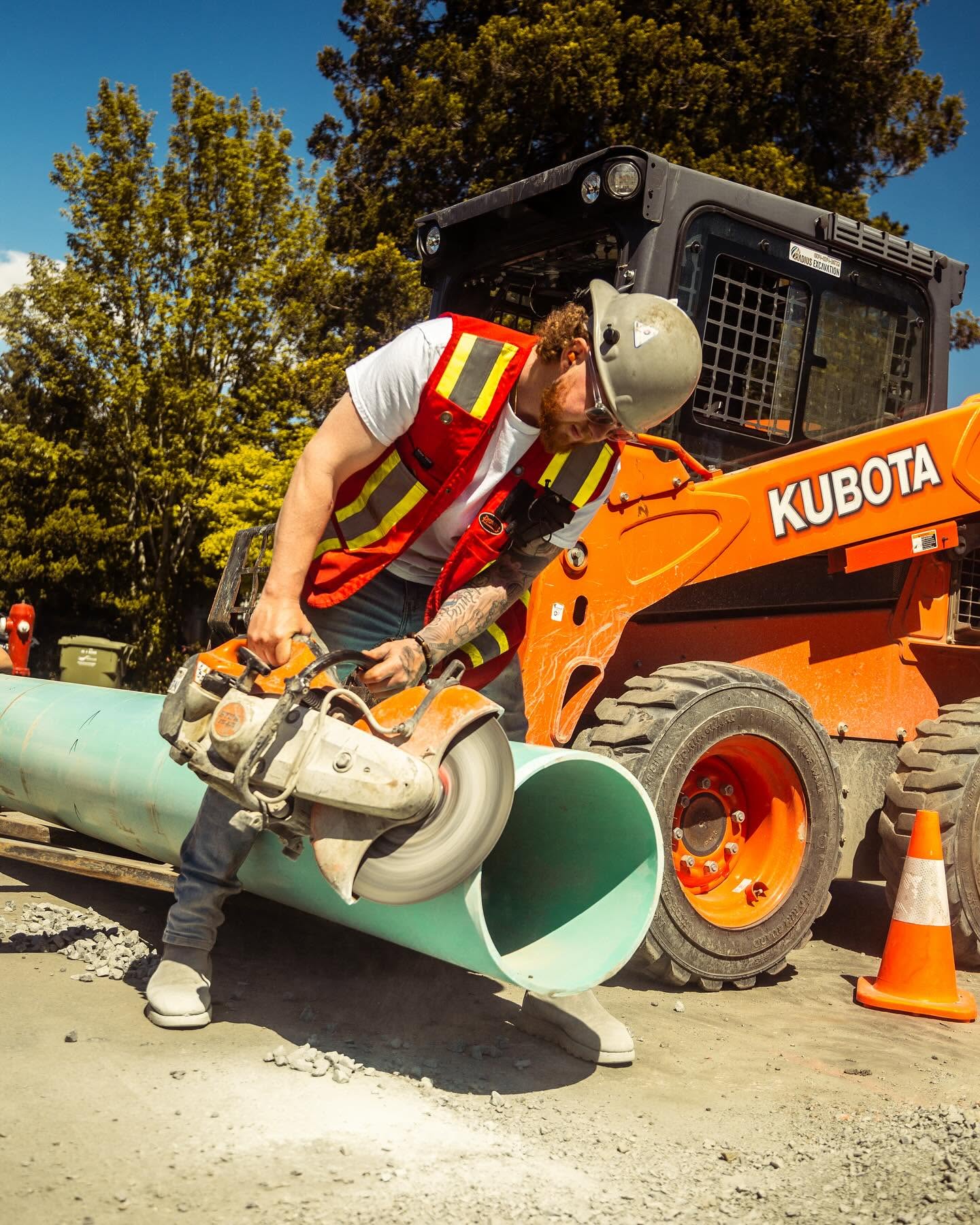 Radius Excavation crew member cutting a large green HDPE storm pipe with a demo saw beside a Kubota skid steer