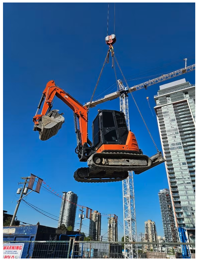 Mini excavator suspended by crane against a downtown tower skyline in Vancouver