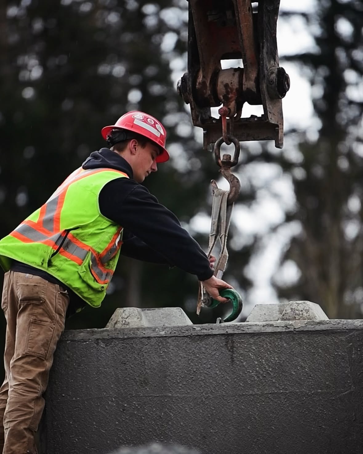 Crew member rigging a chain hook to a precast concrete lock block being lifted by an excavator thumb