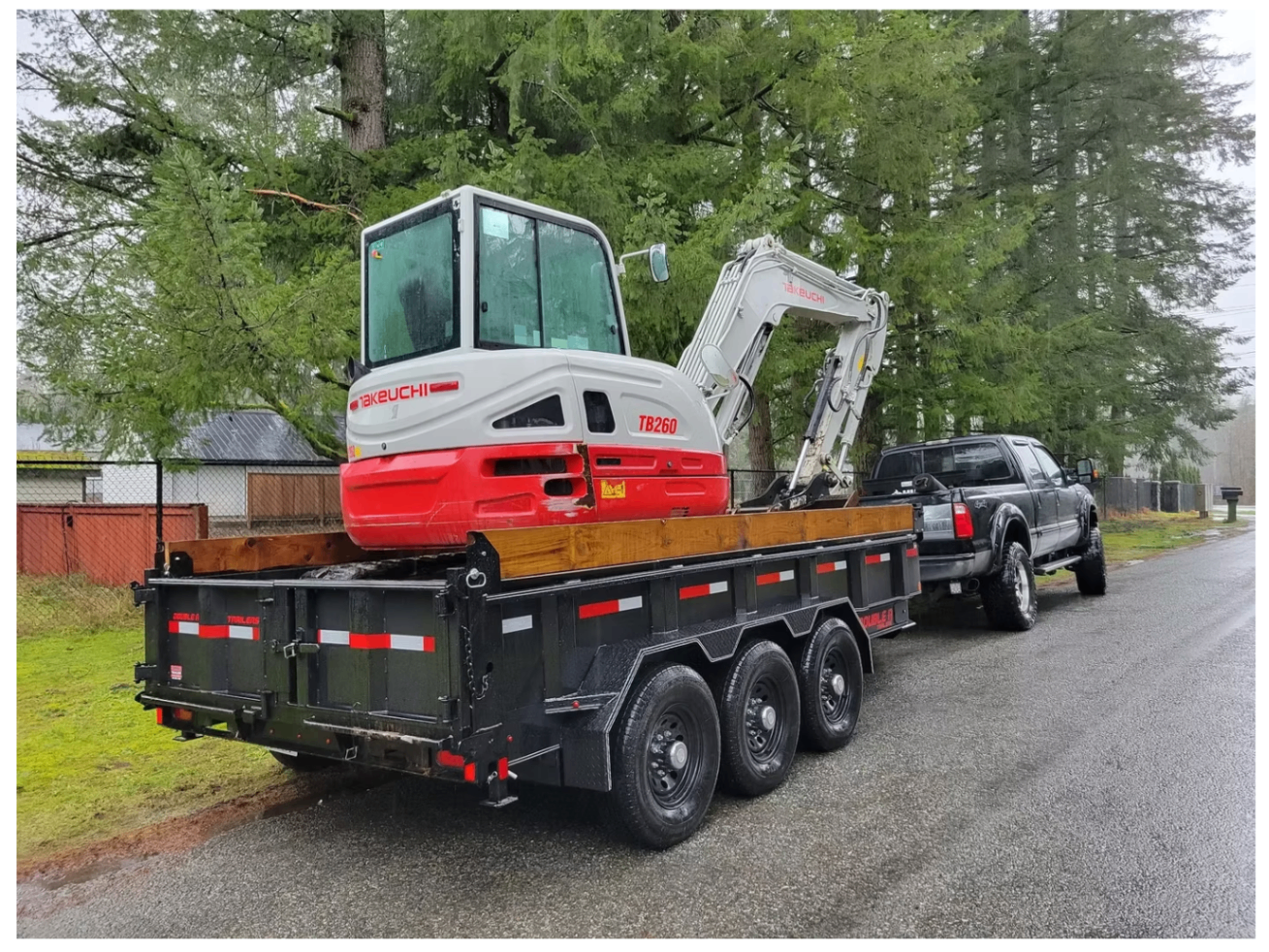 Takeuchi mini excavator loaded on a black dump trailer on a rainy residential street