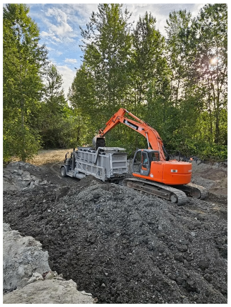 Large excavator loading soil into a dump truck at a wooded excavation site