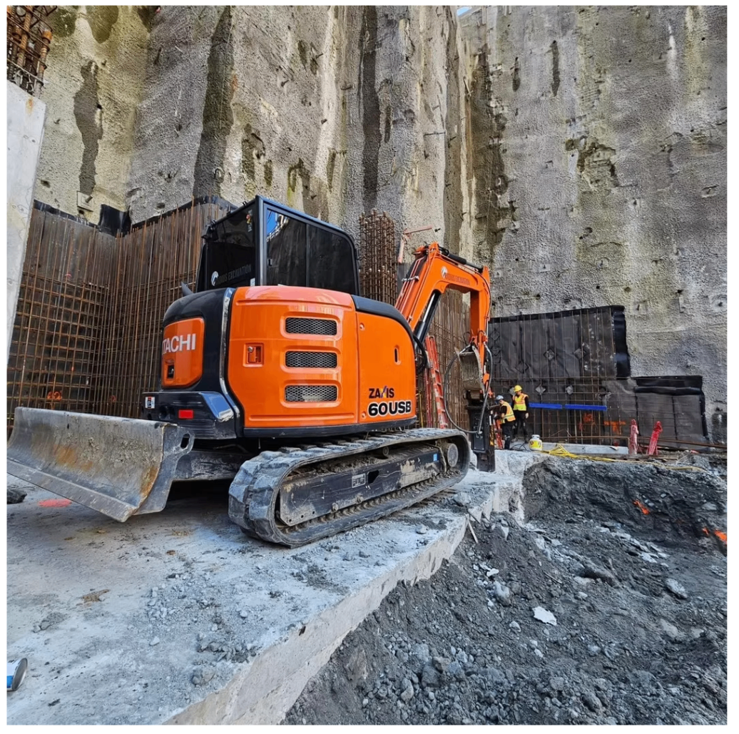 Orange excavator operating at the base of a deep shored excavation with crews nearby