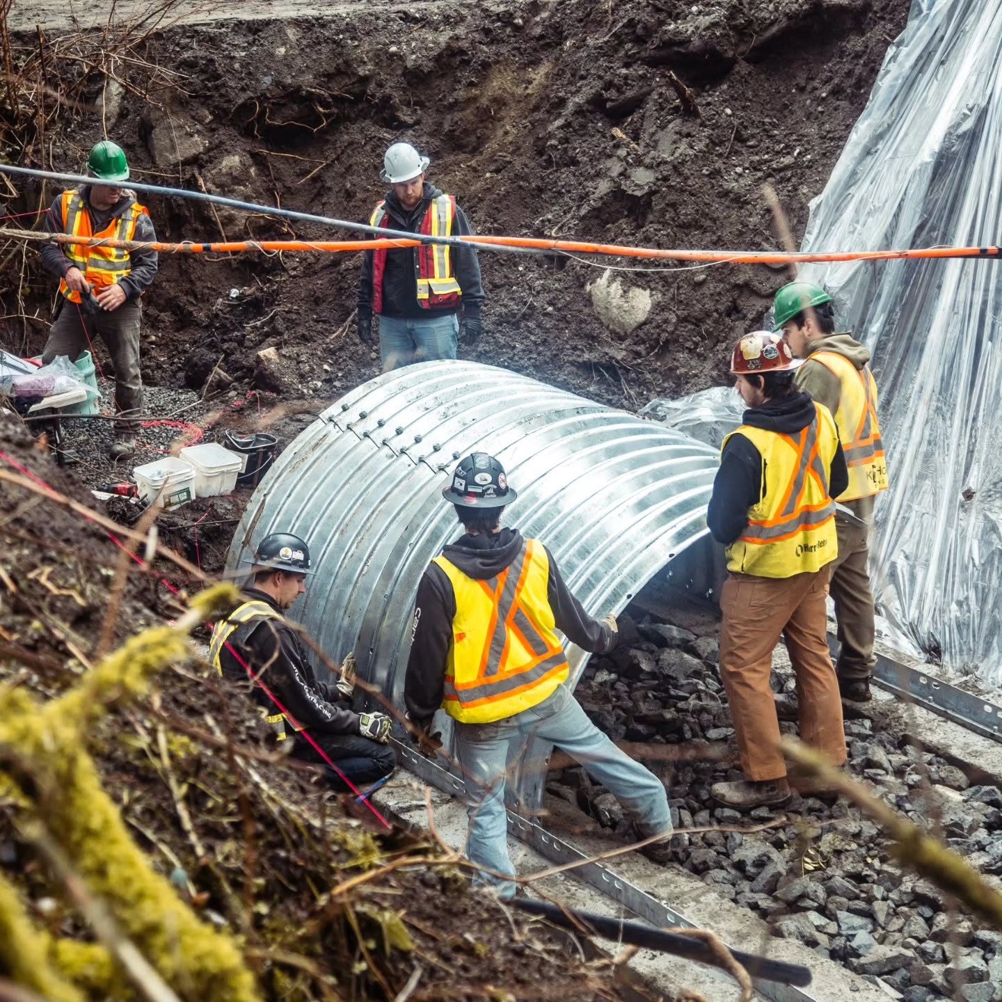 Crew of six installing a large galvanized culvert on a slope repair site with high-visibility vests and hard hats